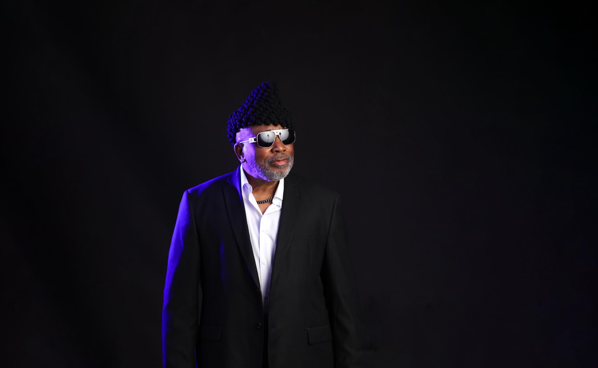 James E. Alexander in a black suit and sunglasses, lit in blue against a dark background