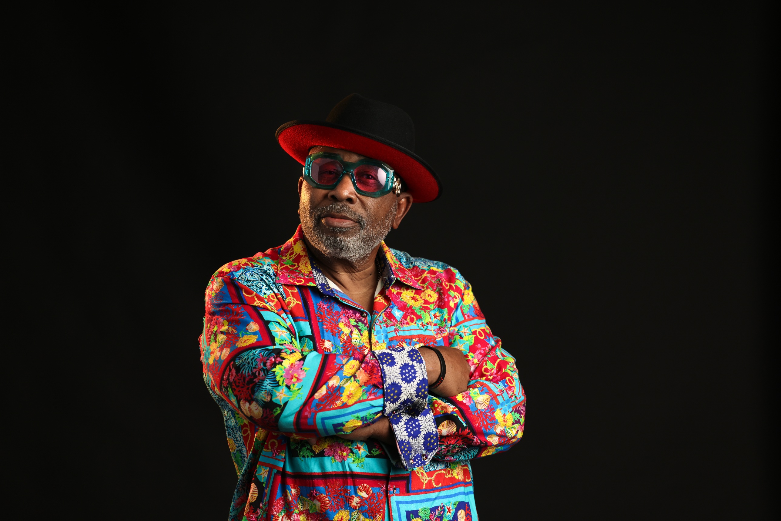 James E. Alexander with arms crossed, wearing a vivid patterned shirt, a red-brim black hat and turquoise glasses